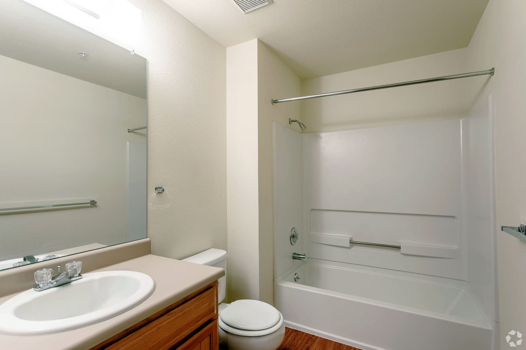 A bathroom with a full tub and shower combination, a wide mirror, a vanity with oak cabinets, and wood-look flooring.