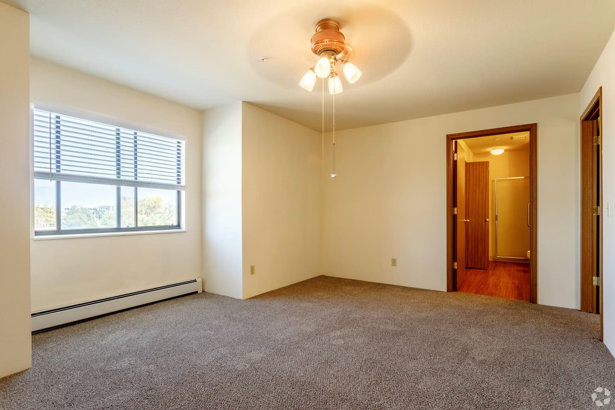 A carpeted bedroom with a large window, a ceiling fan with a light, and a door through to its own bathroom.