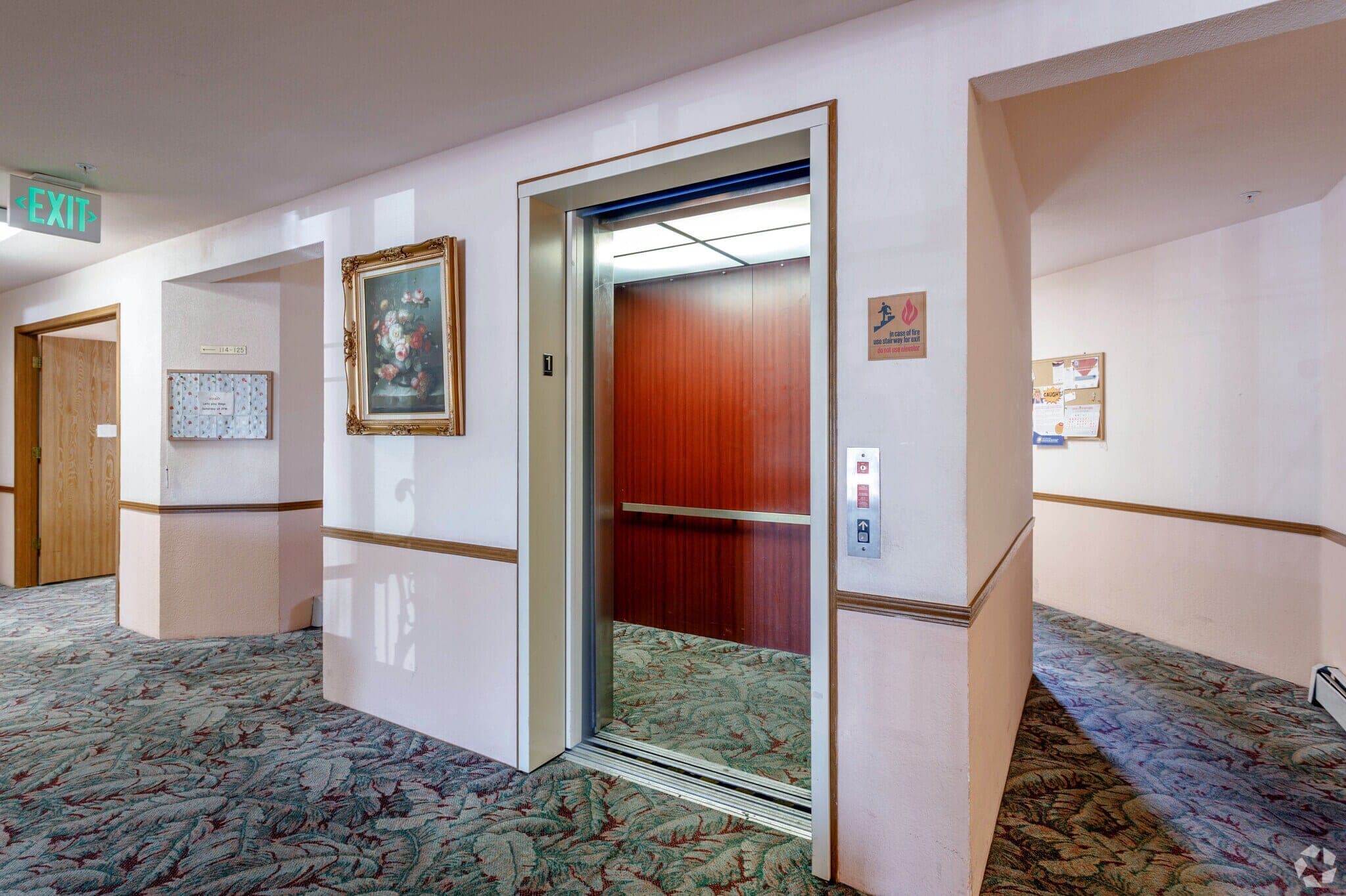 An elevator standing open on a residential floor, beside a resident notice board and a framed painting.