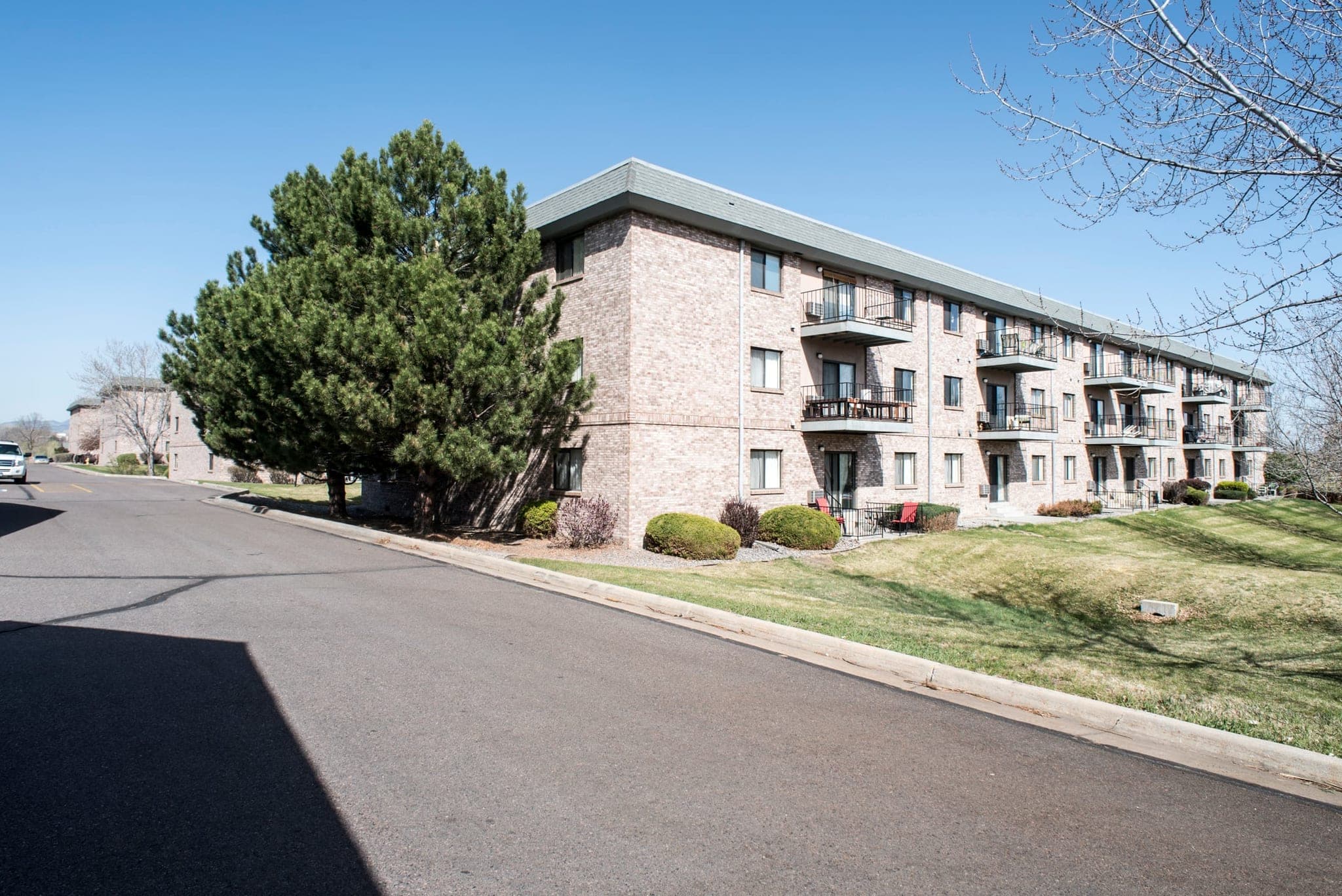 A three-storey stone apartment building at Morningside seen from the parking area, with private balconies on every floor, a mature evergreen beside it and a mown lawn in front.