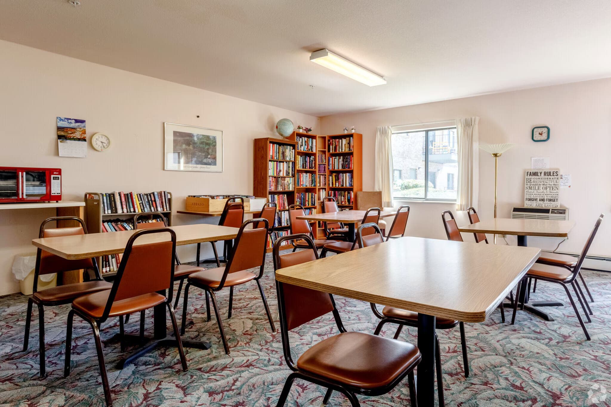 The community library and activity room, with filled bookshelves along one wall, tables and chairs, and a window to the courtyard.