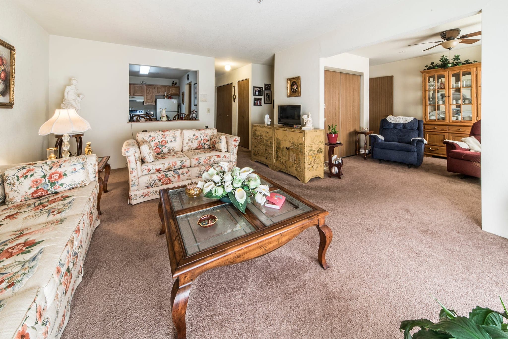 A resident’s furnished living room: two sofas and a coffee table on the carpet, a china cabinet and sideboard along the wall, a ceiling fan above, and the room opening through to the hallway.