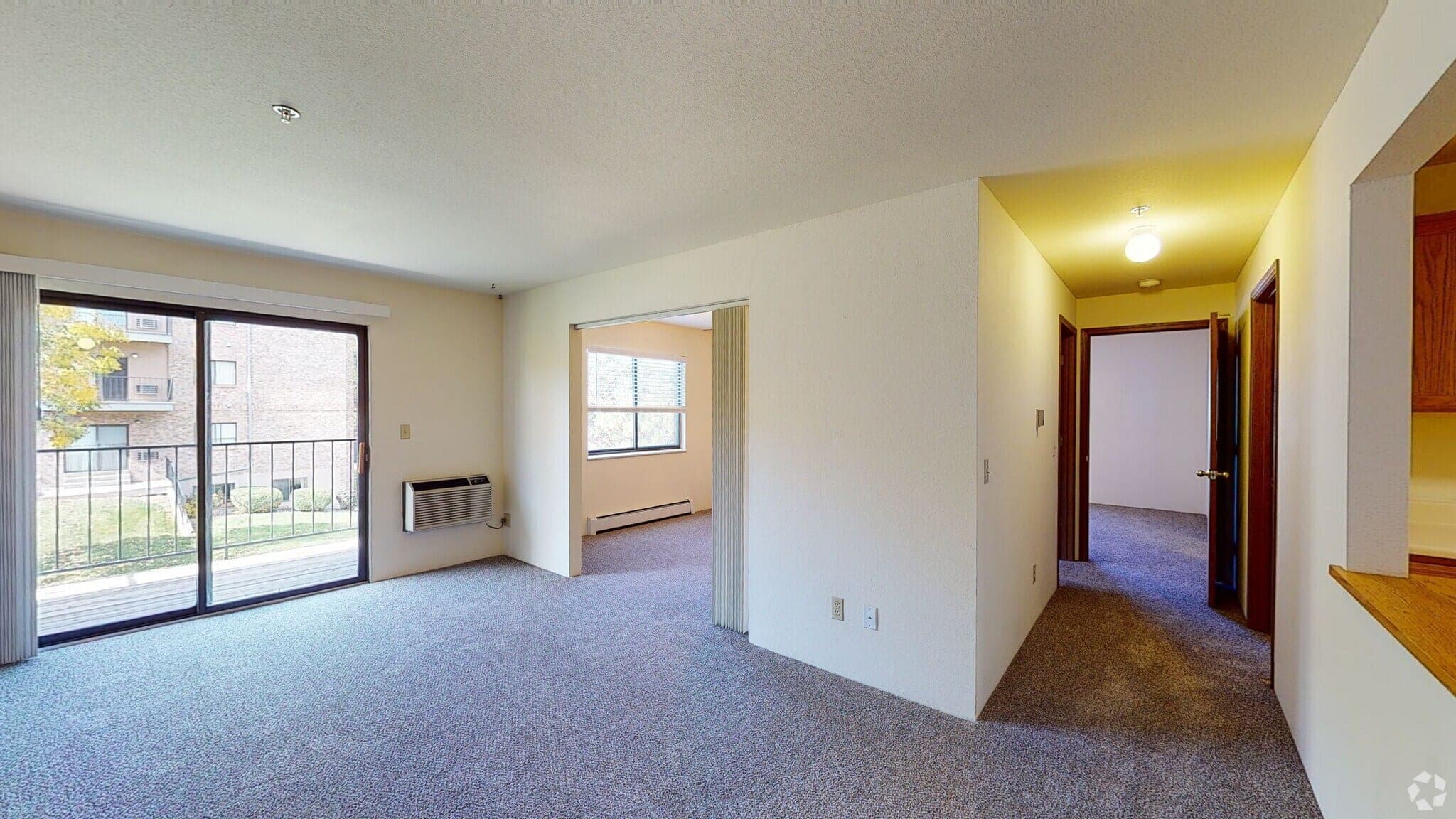 A bright empty living room with a sliding glass door to the balcony, a wall air conditioning unit, and a hallway leading to the bedrooms.
