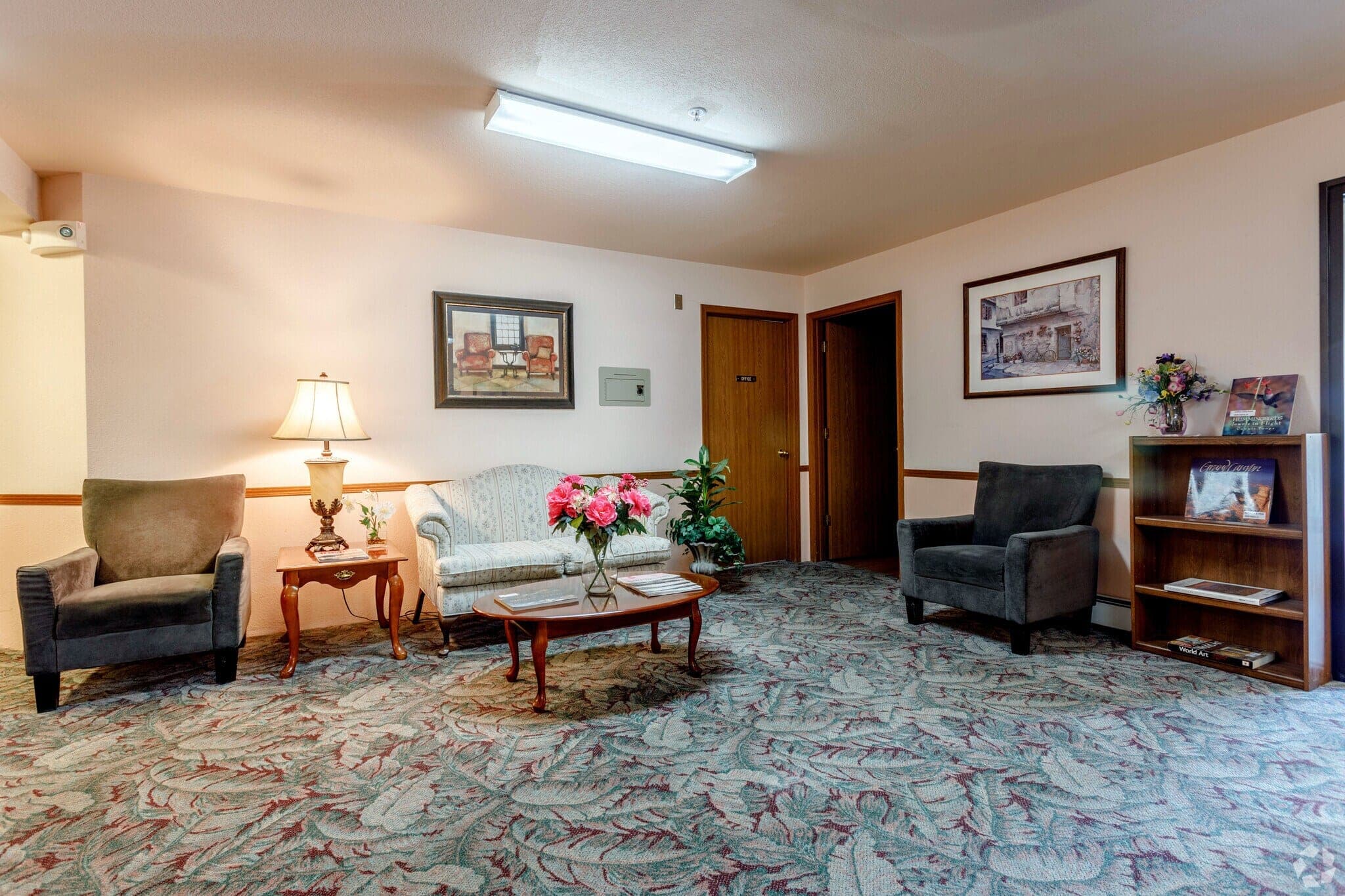 The building lobby, with armchairs and a sofa around a coffee table, a table lamp and the leasing office door.